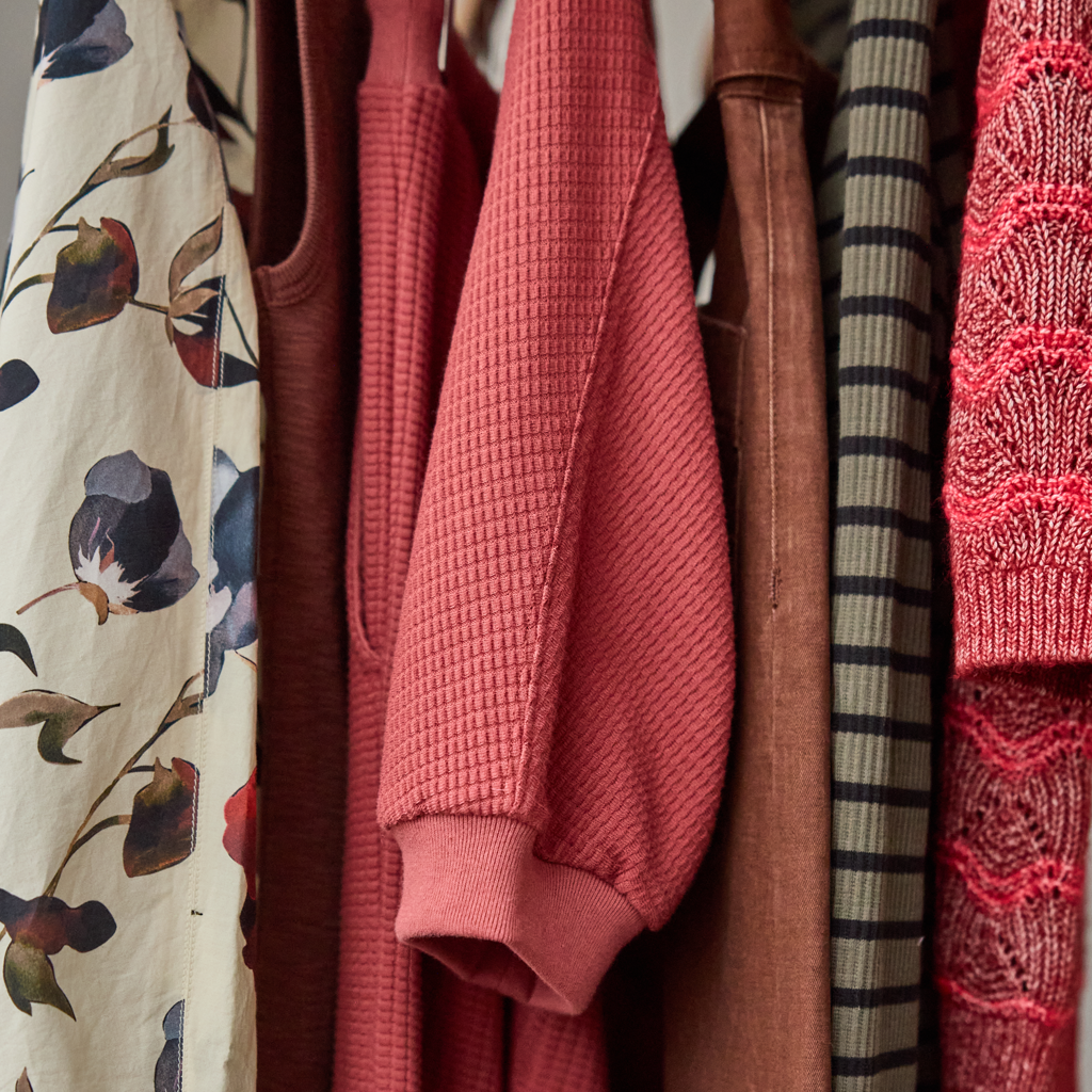 A close-up of hanging garments in a closet, with a dusty rose waffle-knit long-sleeve top centered, surrounded by other clothes including a floral print, brown fabric, striped knit, and coral lace.