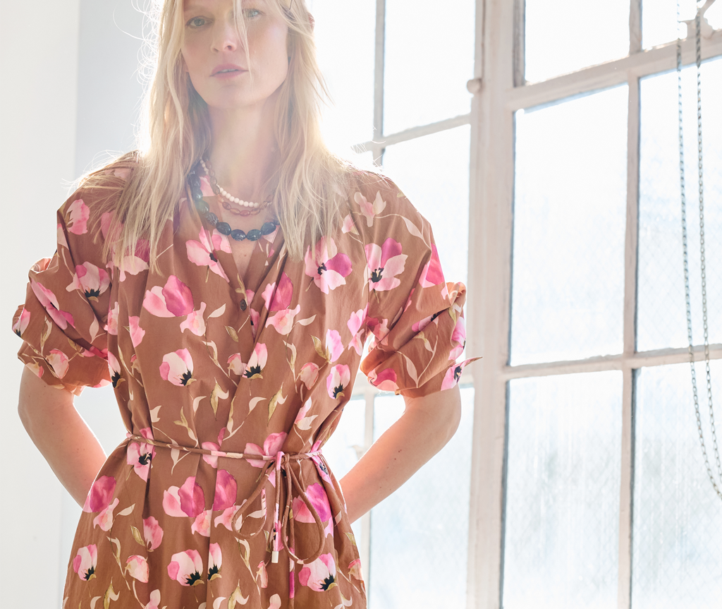 A woman wearing a brown wrap-style dress with a pink and white floral print, standing in front of a bright window.