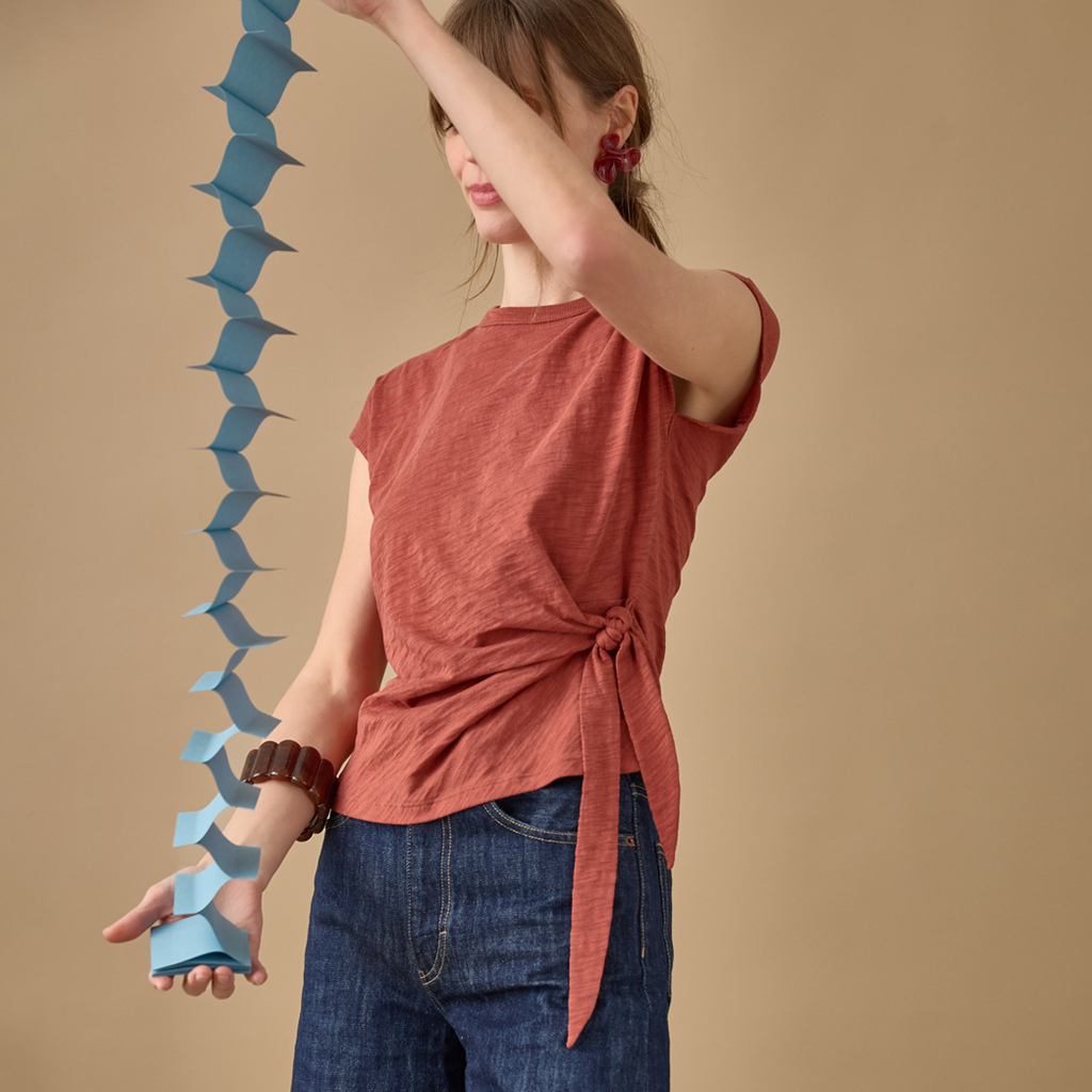 A woman wearing a rust-colored short-sleeve tie-front t-shirt and blue jeans, holding a long blue accordion chain against a solid beige background.