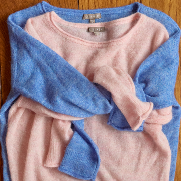 A folded light pink and light blue knit sweater laid flat on a wooden surface, with the sleeves draped over the body of the garment. A neck label reads 'Lilla P.