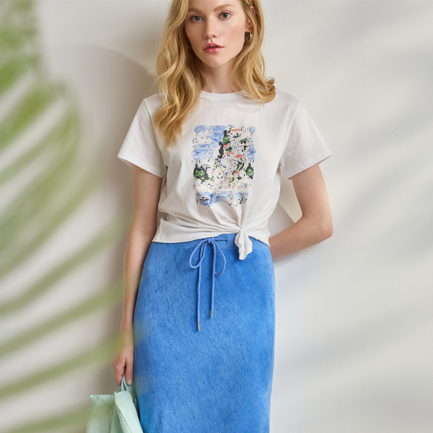 Woman wearing a white t-shirt with a floral design and a blue skirt, holding a green bag.