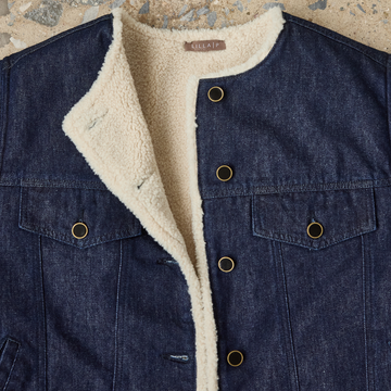 Close-up of a dark indigo denim jacket with a thick cream sherpa lining and brass-tone snap buttons, laid flat on a textured stone surface. The brand name 'GALRAFT' is visible on the inner neck label.