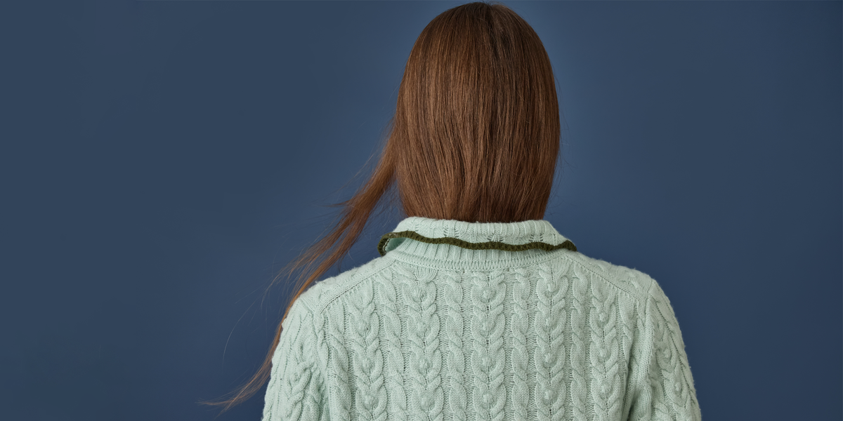 A woman viewed from behind wearing a pale sage green cable-knit sweater against a solid dark blue background.