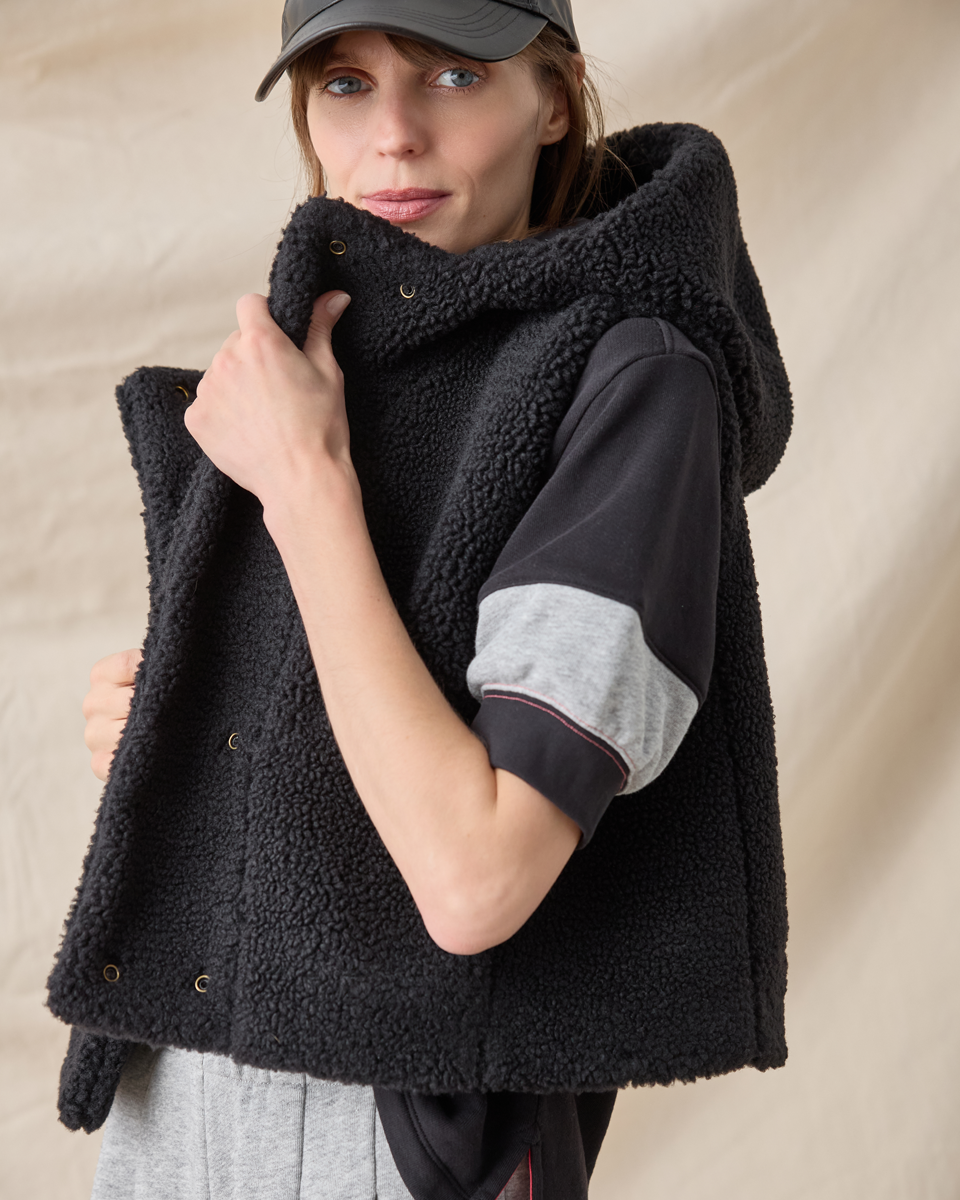 A woman wearing a black sherpa fleece hooded vest over a black and gray color-blocked t-shirt, paired with a dark gray baseball cap, against a neutral beige fabric backdrop.