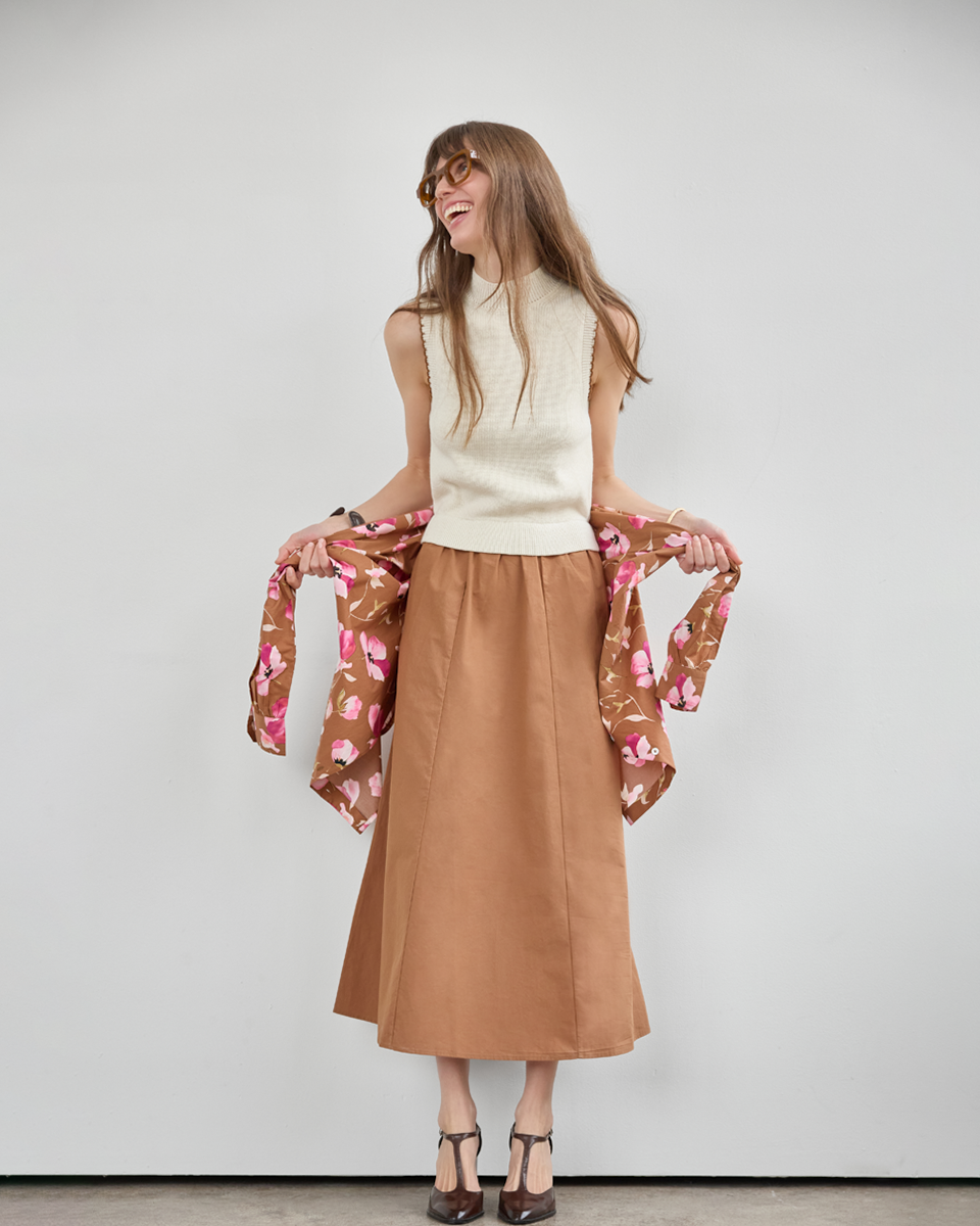 A woman stands against a light gray studio wall wearing a cream sleeveless knit top, a brown midi skirt, and holding a brown floral cardigan. She is wearing brown strappy heels.