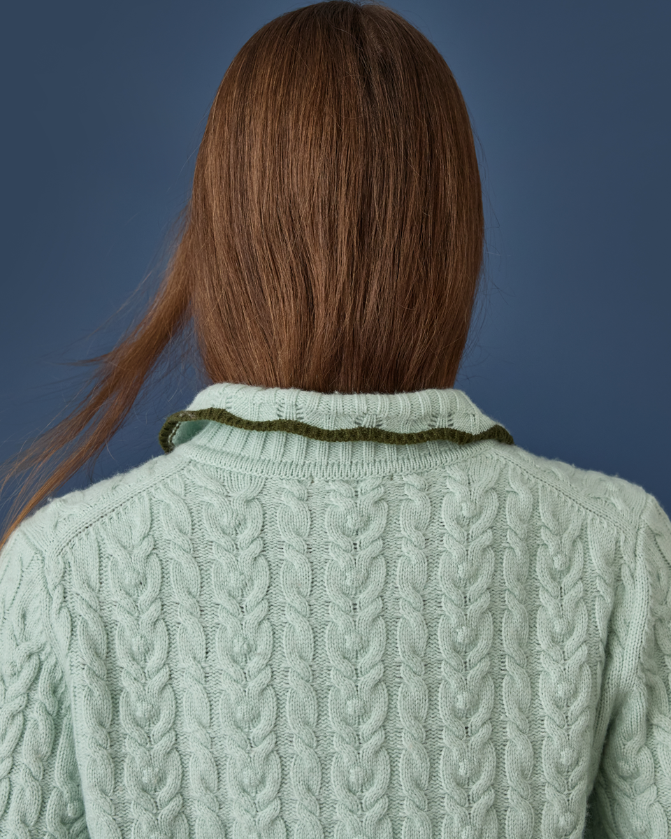 Back view of a person with long brown hair wearing a pale sage green cable-knit sweater with a contrasting dark olive green collar trim, set against a solid dark blue background.