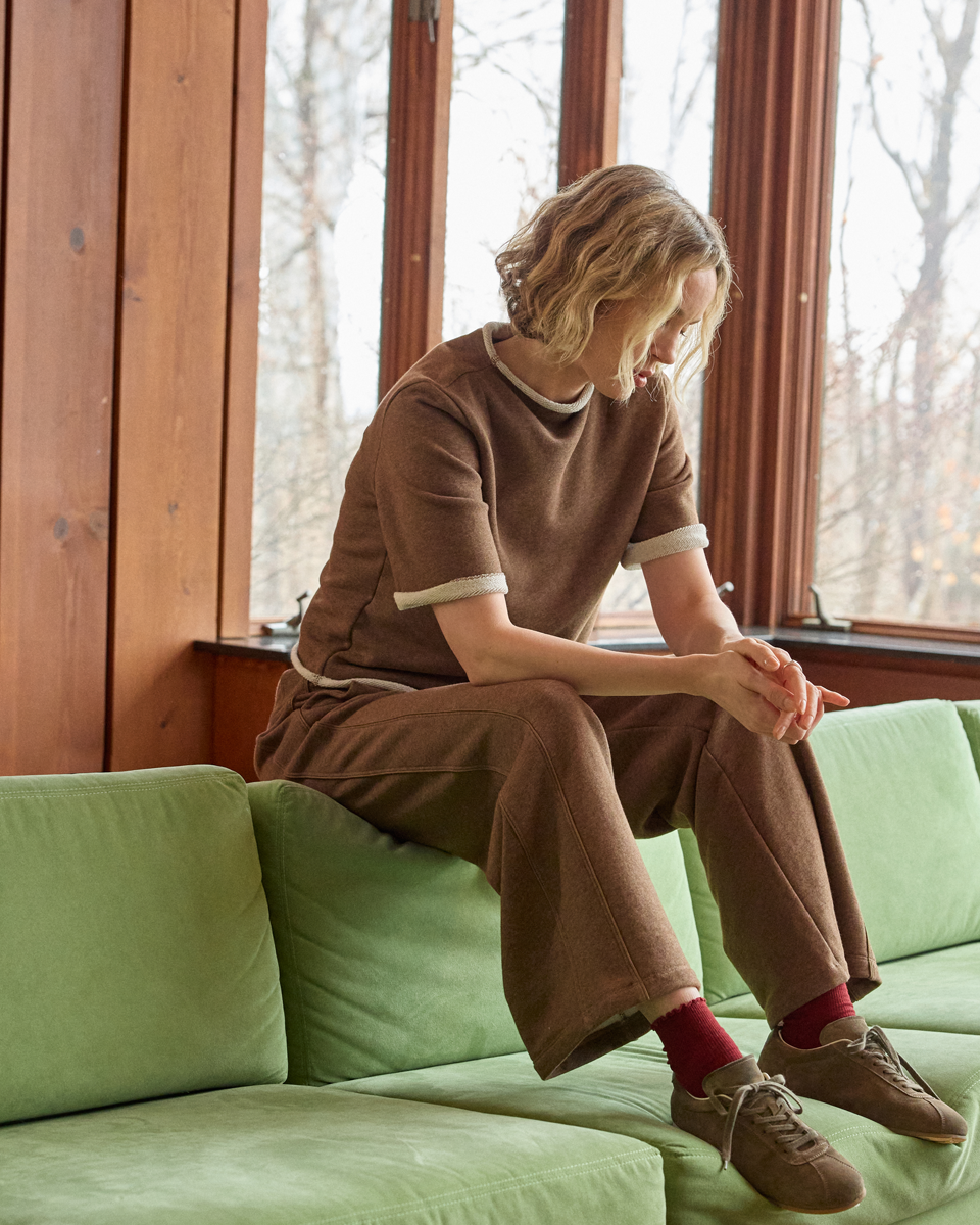 A woman sits on a light green sofa in a room with wood-paneled walls and large windows. She is wearing a matching brown knit short-sleeve top and wide-leg pants with white ribbed trim, red socks, and brown sneakers.