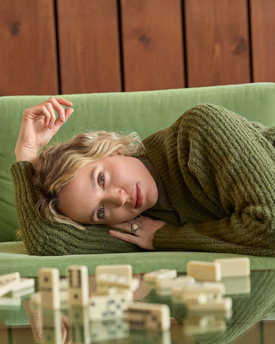 A woman with blonde hair lies on a green couch, wrapped in a chunky olive green knit sweater. In the foreground, wooden dominoes are scattered on a glass table against a dark wood-paneled wall.