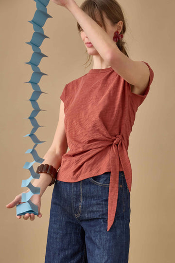 A woman modeling a rust-colored short-sleeve top with a side tie detail, paired with dark blue wide-leg jeans, against a solid beige background.