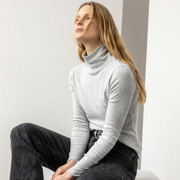 A blonde model sits on a white box in front of a white wall wearing black jeans and a gray fitted turtleneck. The turtleneck is style #pa1278 in heather gray